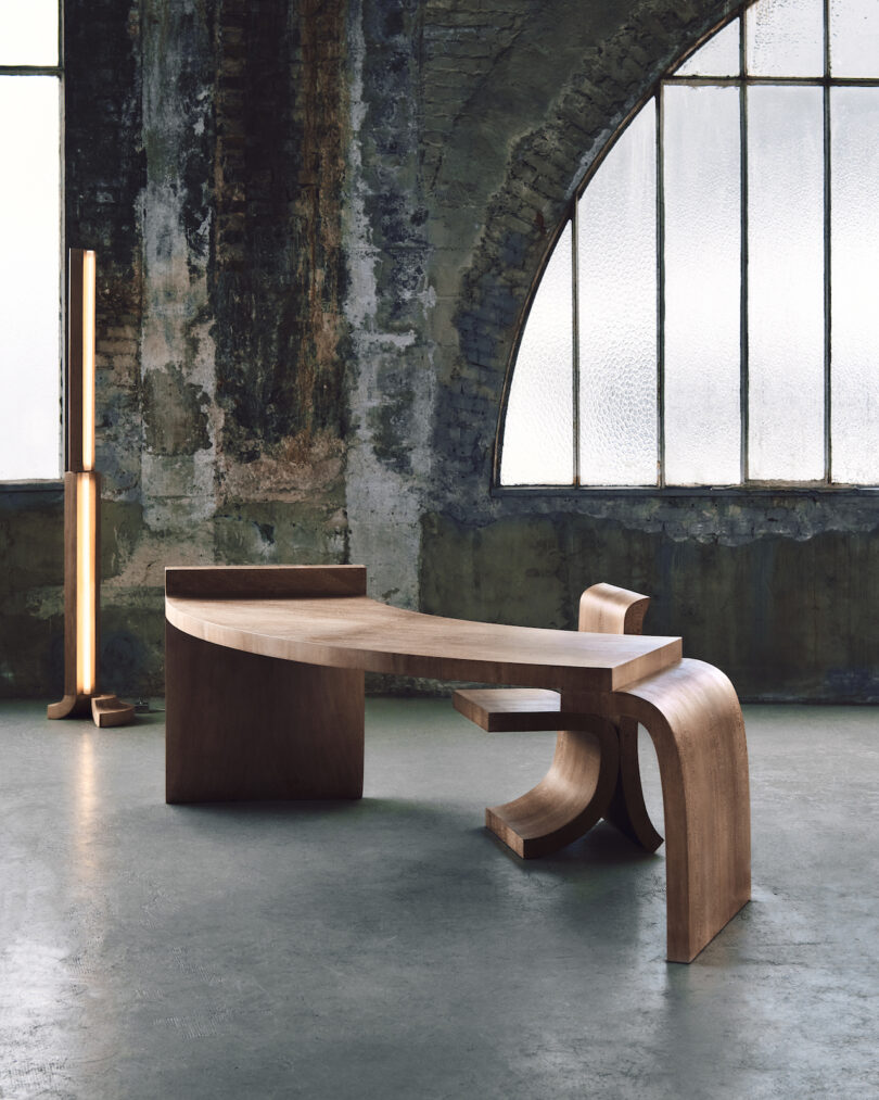 A modern, uniquely-shaped wooden desk and chair set against a rustic industrial backdrop with large, arched windows and a vertical LED light fixture.