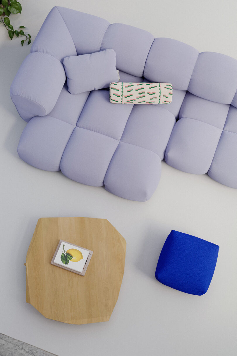 A lavender sectional sofa with pillows, a blue ottoman, and a wooden coffee table with a book and a lemon illustration on it, arranged in a minimalist living room