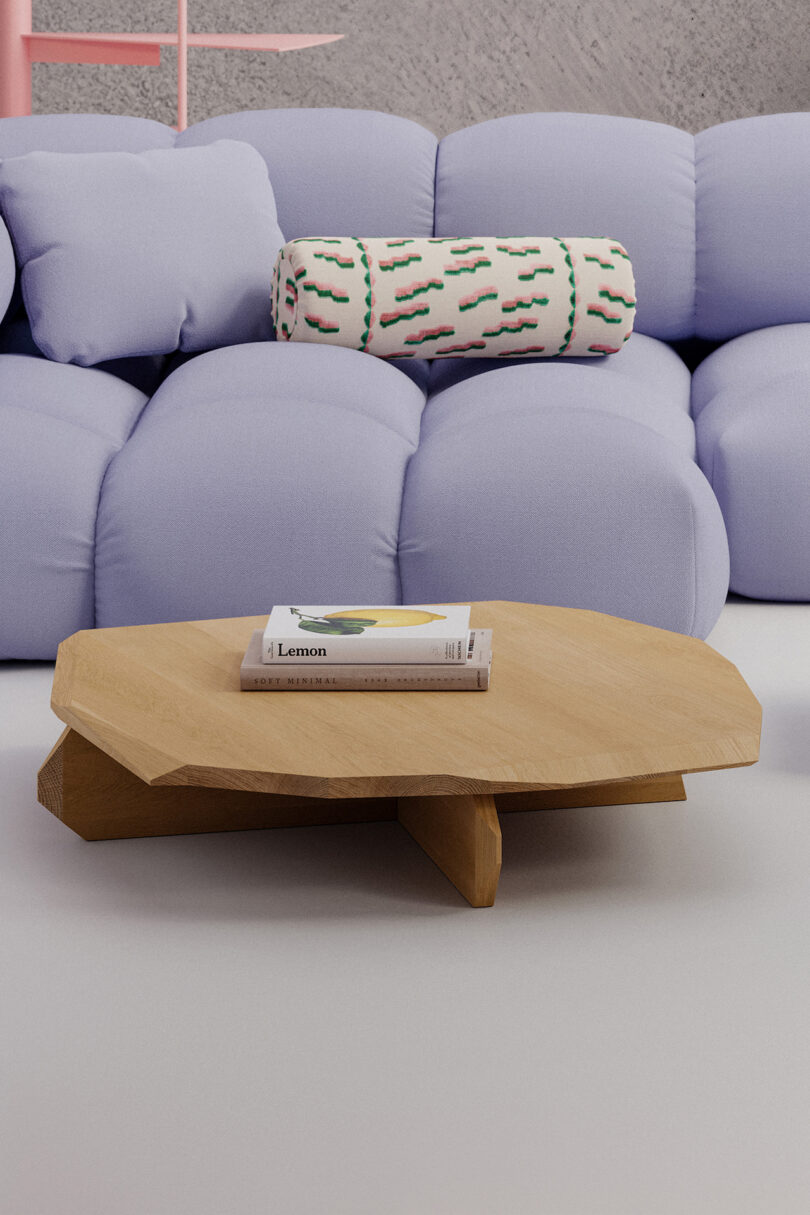 A low wooden coffee table with a book titled "Lemon" is in front of a pastel blue sofa with a cylindrical patterned cushion in a minimalist living room