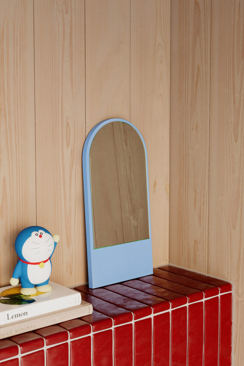 Small blue mirror on red tiled surface next to books and a figurine of a blue robotic cat. Background consists of light wooden paneling