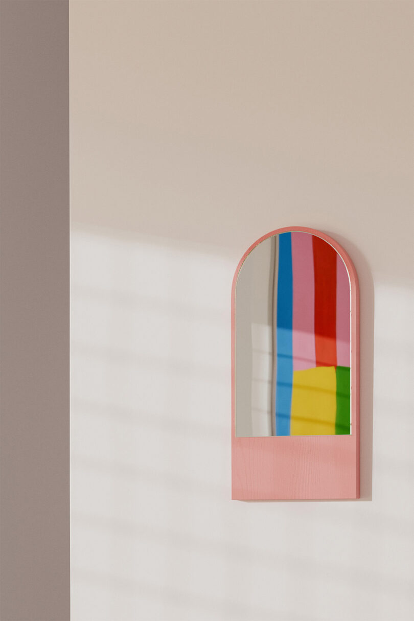 A small arched mirror with a pink frame reflects colorful rectangles under soft light on a beige wall