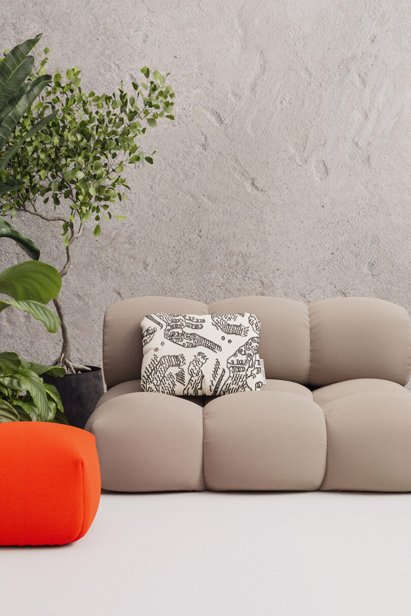 A beige modular sofa with a patterned cushion is placed against a gray textured wall, accompanied by a tall potted plant. In the foreground, there is a bright orange pouf