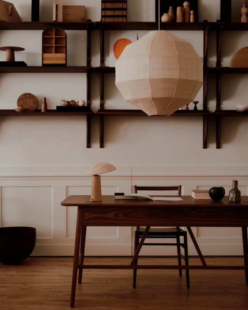 A minimalist office setup with a wooden desk and chair, overhead paper lantern, and shelves displaying various wooden decor items.