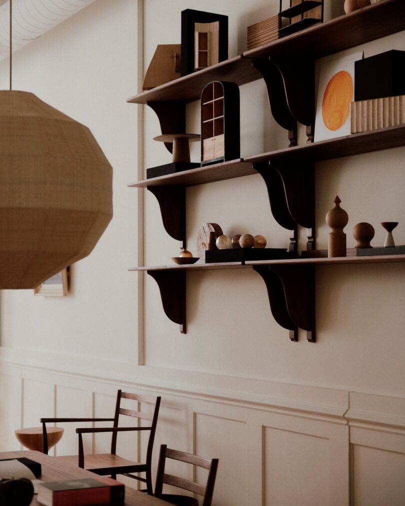 A wooden shelf with geometric decorations and small sculptures, mounted on a cream-colored wall above a wooden table and chairs. A large pendant light hangs from the ceiling.