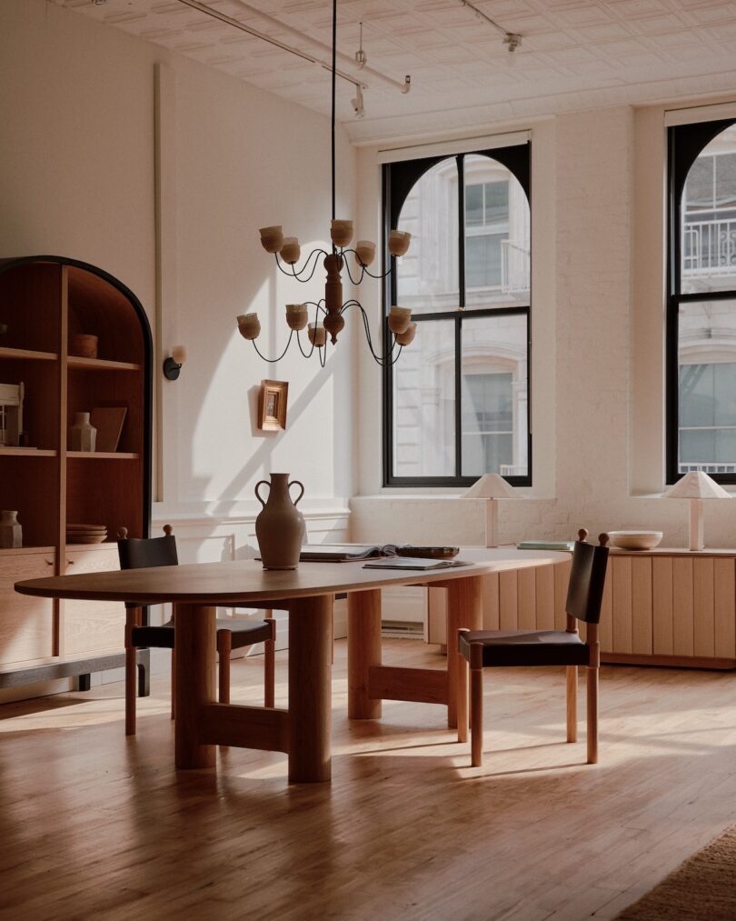 A well-lit dining room features a wooden table with a vase, chairs, a modern chandelier, large arched windows, and a wooden shelf unit against a white brick wall.