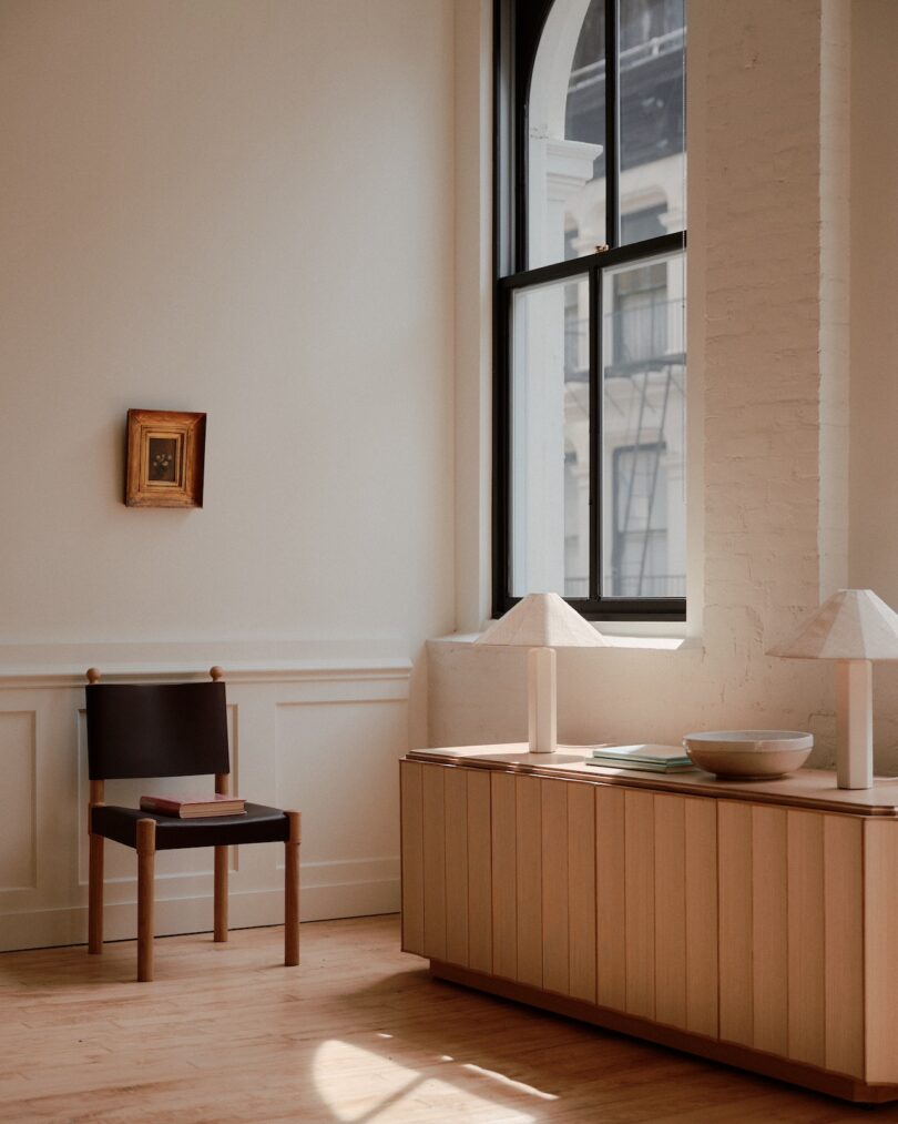 Minimalist room with chair, small framed picture on wall, large window, and sideboard with lamps, books, and bowl. Soft natural light fills the space, highlighting its neutral tones and simple decor.