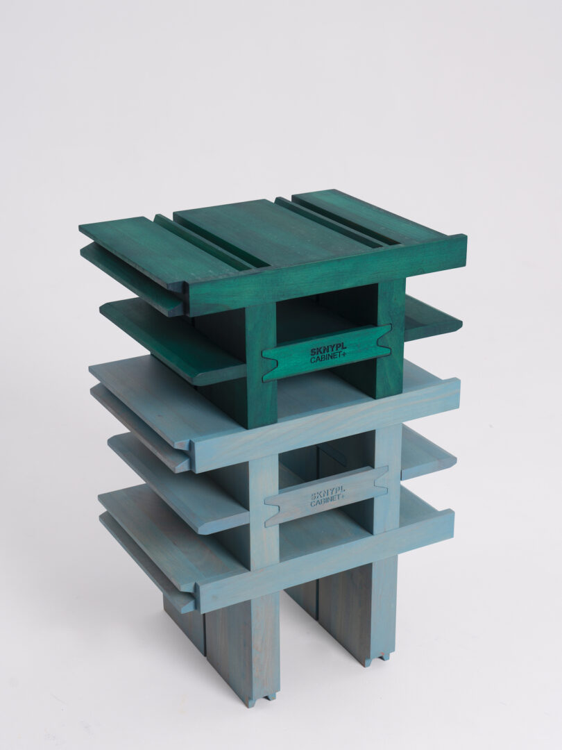 A small, multi-tiered rectangular shelf unit made of green and blue wood, with the words "Skopyta Cabinet" printed on each tier. The design features open sides and a modular construction