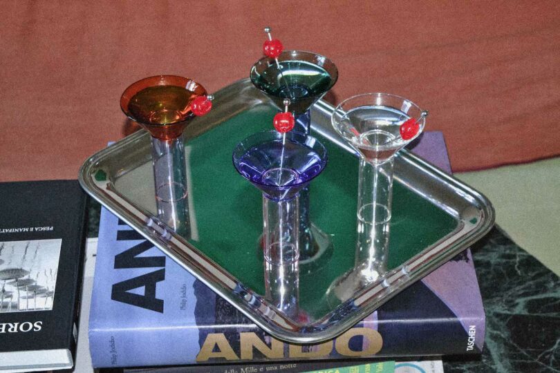 four colorful, stemmed cocktail glasses with red cherries on picks are arranged on a metal tray, placed on top of a stack of books.