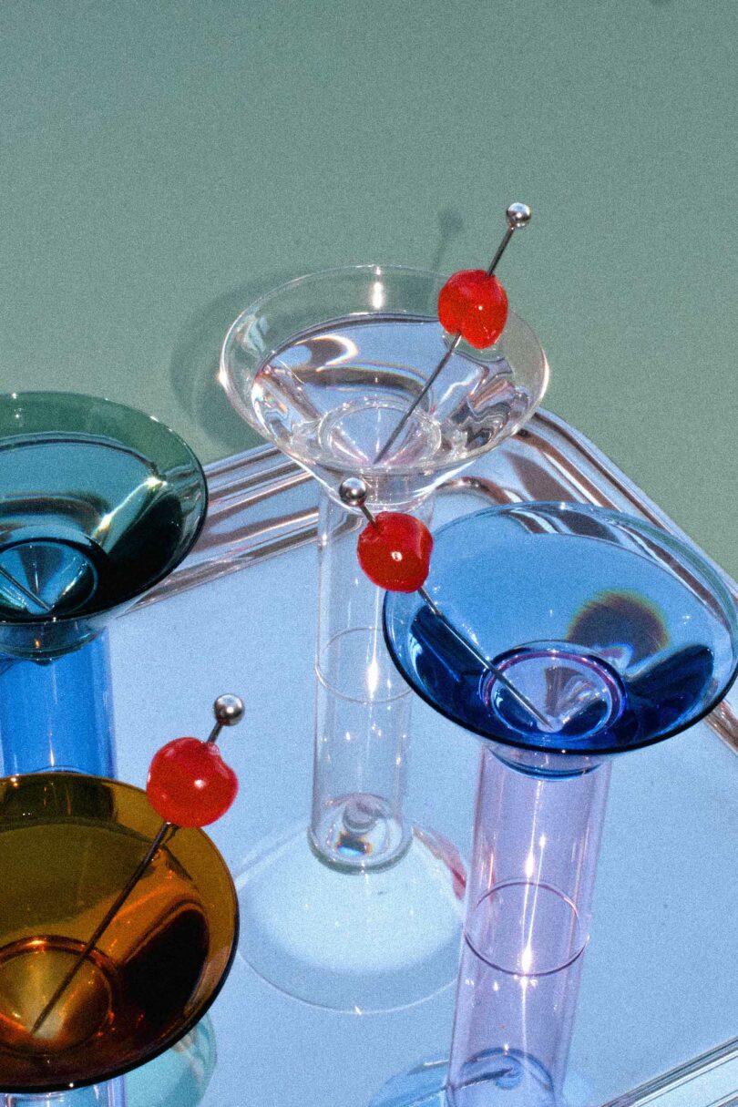 Four colorful cocktail glasses with red cherry garnishes on picks, placed on a reflective surface against a muted green background