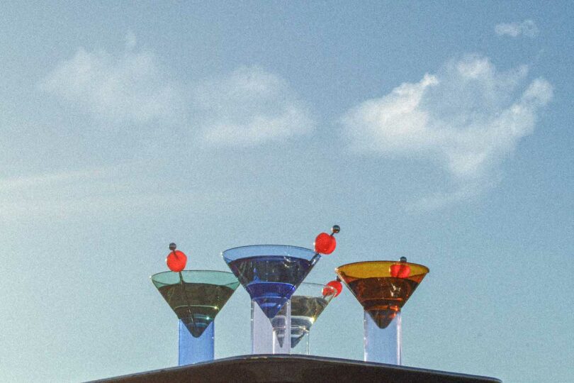 Three colorful cocktails with garnish are arranged in a row against a clear blue sky with a few clouds