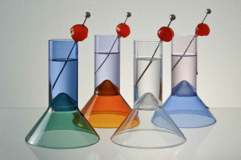 Four colored liquid-filled glasses with long metal stirrers each topped with a red cherry