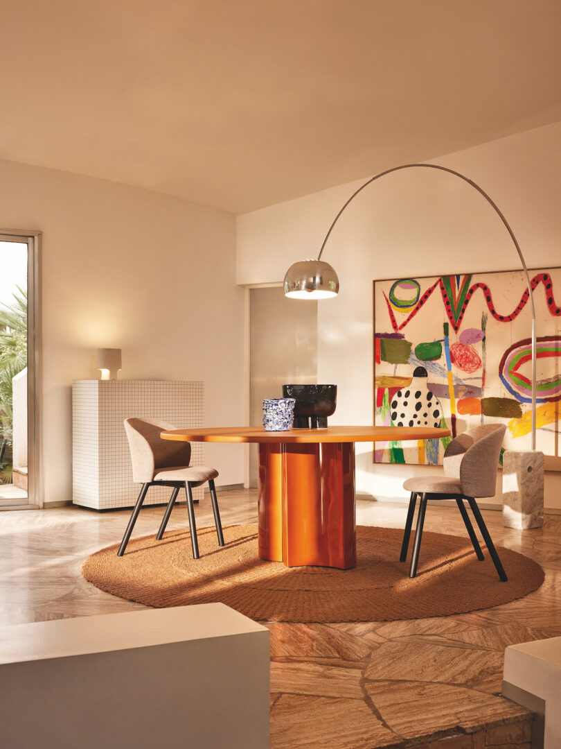 Modern dining room with two upholstered chairs around an orange table, under a curved floor lamp. The room features abstract wall art, a small circular rug, and natural light from a window