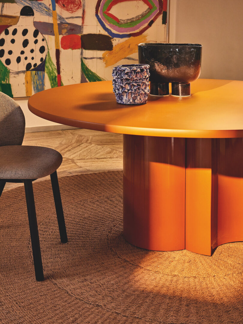 A round orange table with two decorative vases. A gray chair is on the left, and a colorful abstract painting is on the wall in the background. The floor has a woven rug