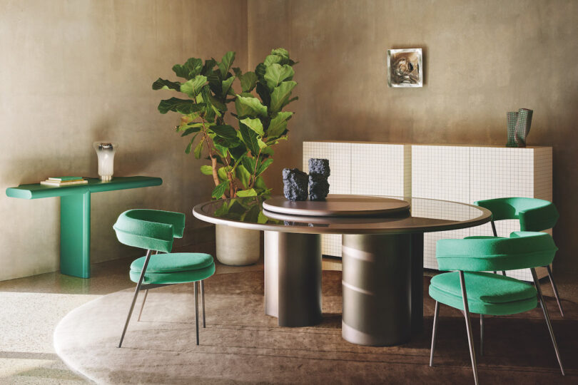 A modern dining room with a round table, green chairs, a potted plant, a console table with decor, and an art piece on the wall