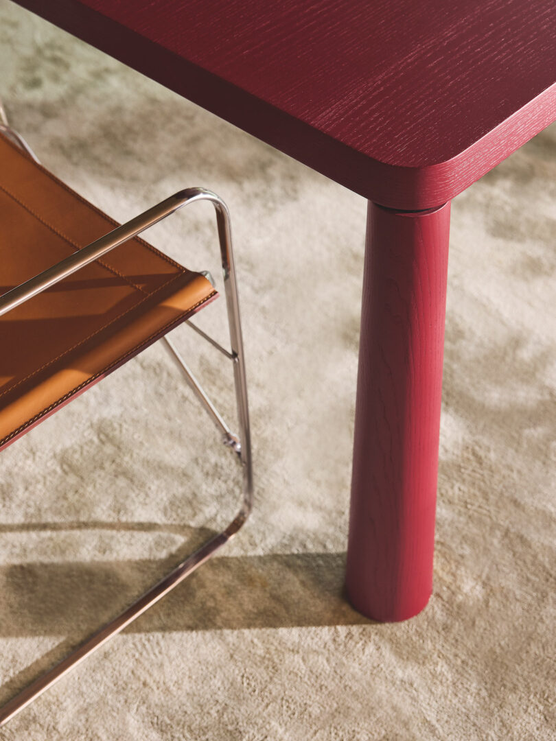 A red wooden table leg is shown next to a tan leather and metal chair on a light-colored carpet