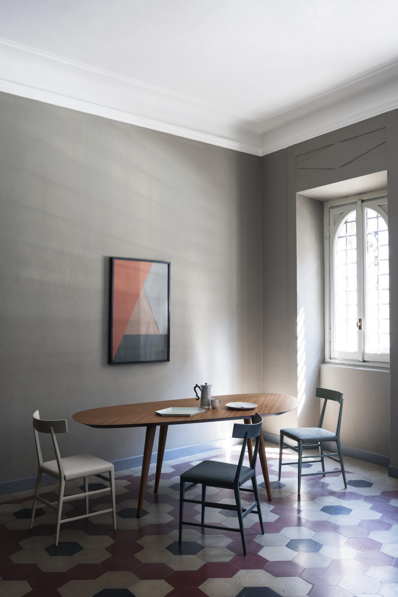 A minimalist dining room with a wooden table, three chairs, a teapot, scattered papers, a geometric painting on a gray wall, and natural light streaming in through a window with white blinds