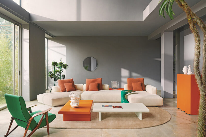 Modern living room with beige sectional sofa, orange and green accents, large windows, light gray walls, indoor plants, unique coffee tables, and contemporary decor