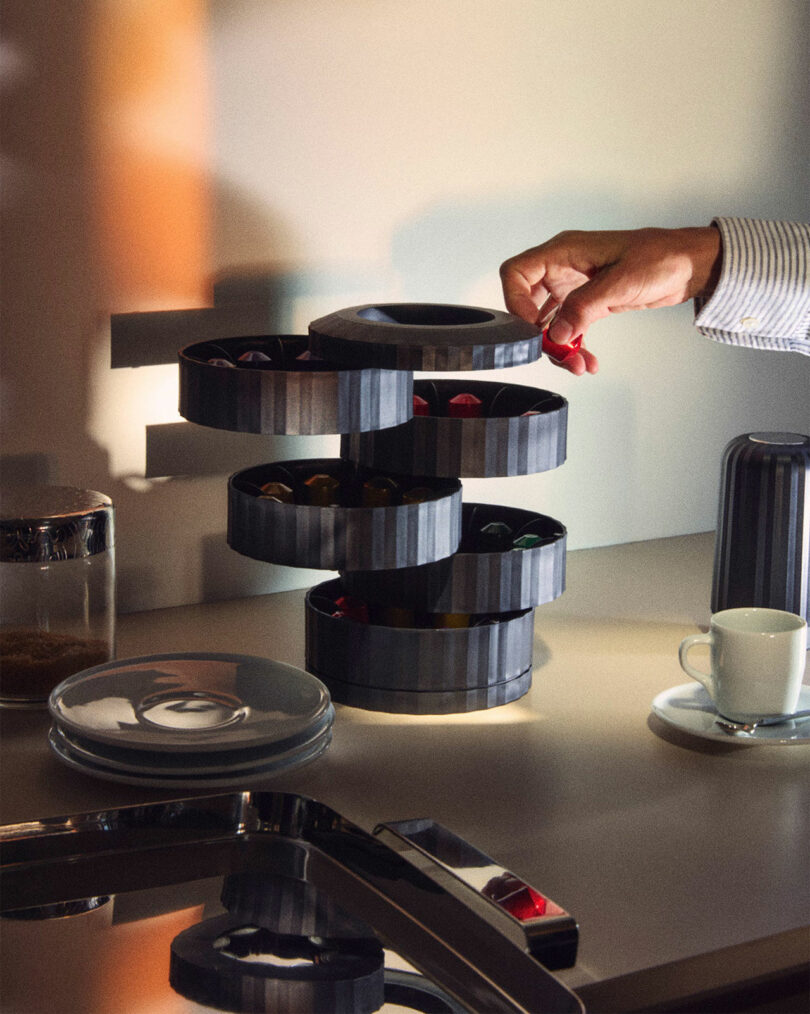 A hand reaching for chocolates in a multi-tiered rotating dispenser on a countertop, with plates, a cup, and a coffee machine nearby.