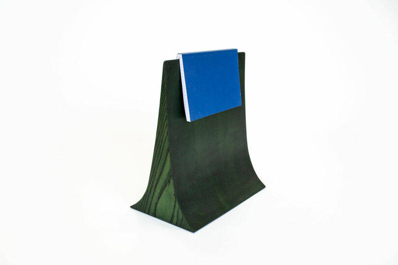 A blue book rests on a green, wave-shaped wooden stand against a white background.