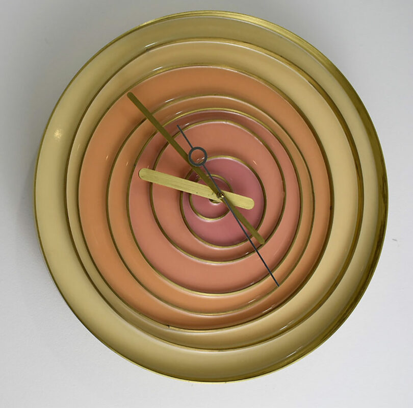 A wall clock with concentric circles in shades of gold, pink, and peach, featuring gold hour and minute hands.