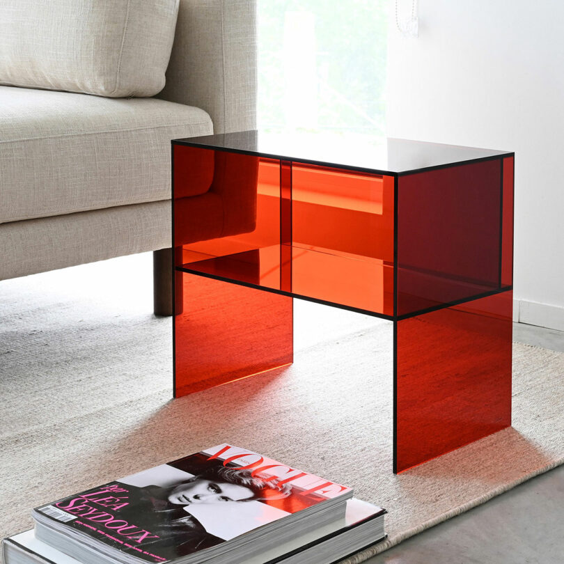 A red transparent glass table beside a beige sofa on a light rug, with a fashion magazine on the floor.