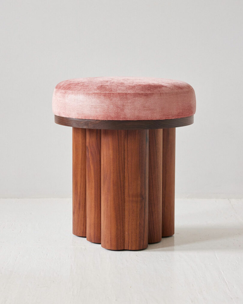 Pink velvet cushioned seat with a cylindrical, multi-legged wooden base on a white floor.