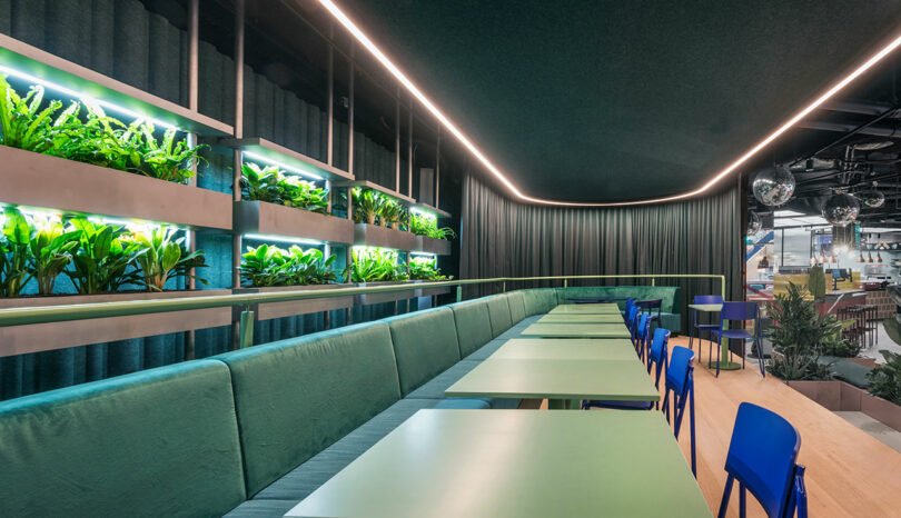 Modern interior with green tables, blue chairs, and wall planters. Ceiling lighting creates a sleek atmosphere.