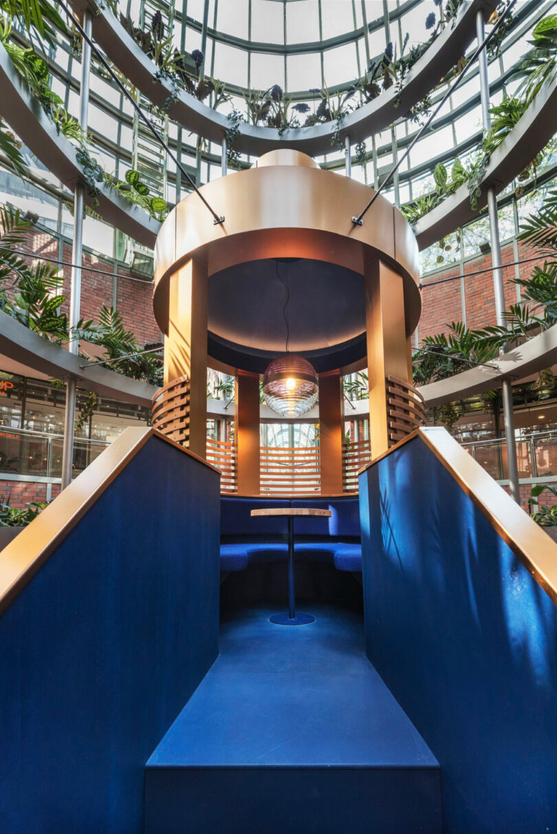 Circular indoor seating area with a blue interior, surrounded by large windows and greenery. A spherical light fixture hangs from the ceiling.