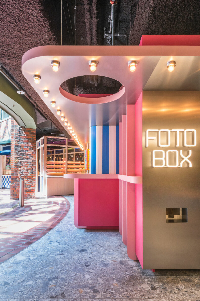 Colorful photobooth area with pink and blue panels, circular cut-out ceiling, and illuminated "Foto Box" sign, surrounded by exposed brick and vibrant lighting.