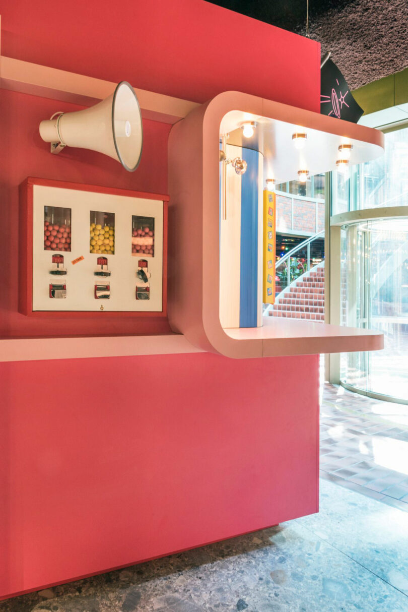 A retro-style pink wall display with a megaphone, photos, and a curved shelf inside a bright, modern building.