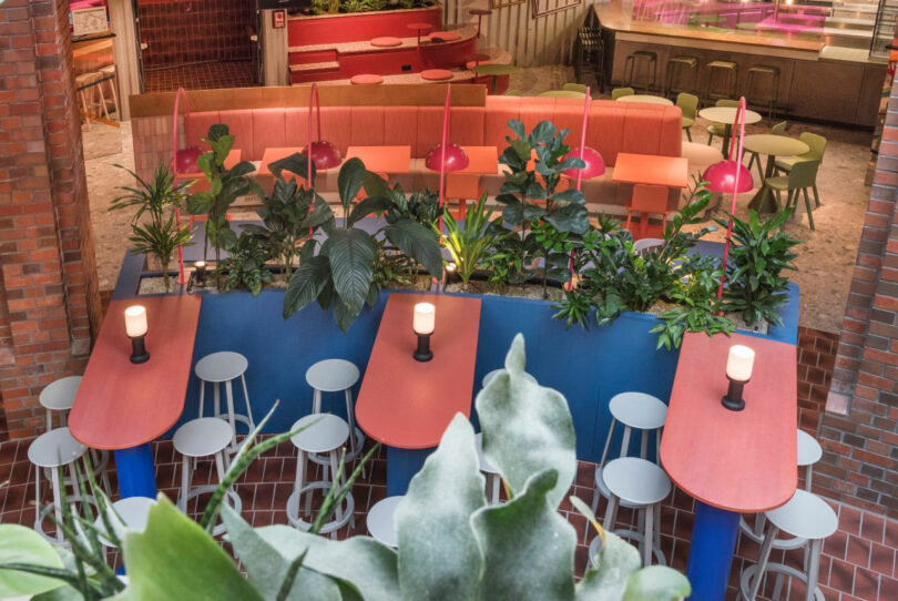 Colorful indoor seating area with red tables, white stools, and green plants. Candles are placed on the tables, surrounded by ambient lighting and brick walls.