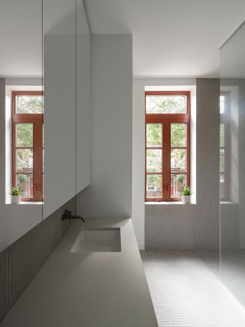 Minimalist bathroom with a gray countertop, sink, and mirrored cabinets. Two windows with wooden frames let in natural light. Small potted plants are on the windowsills.