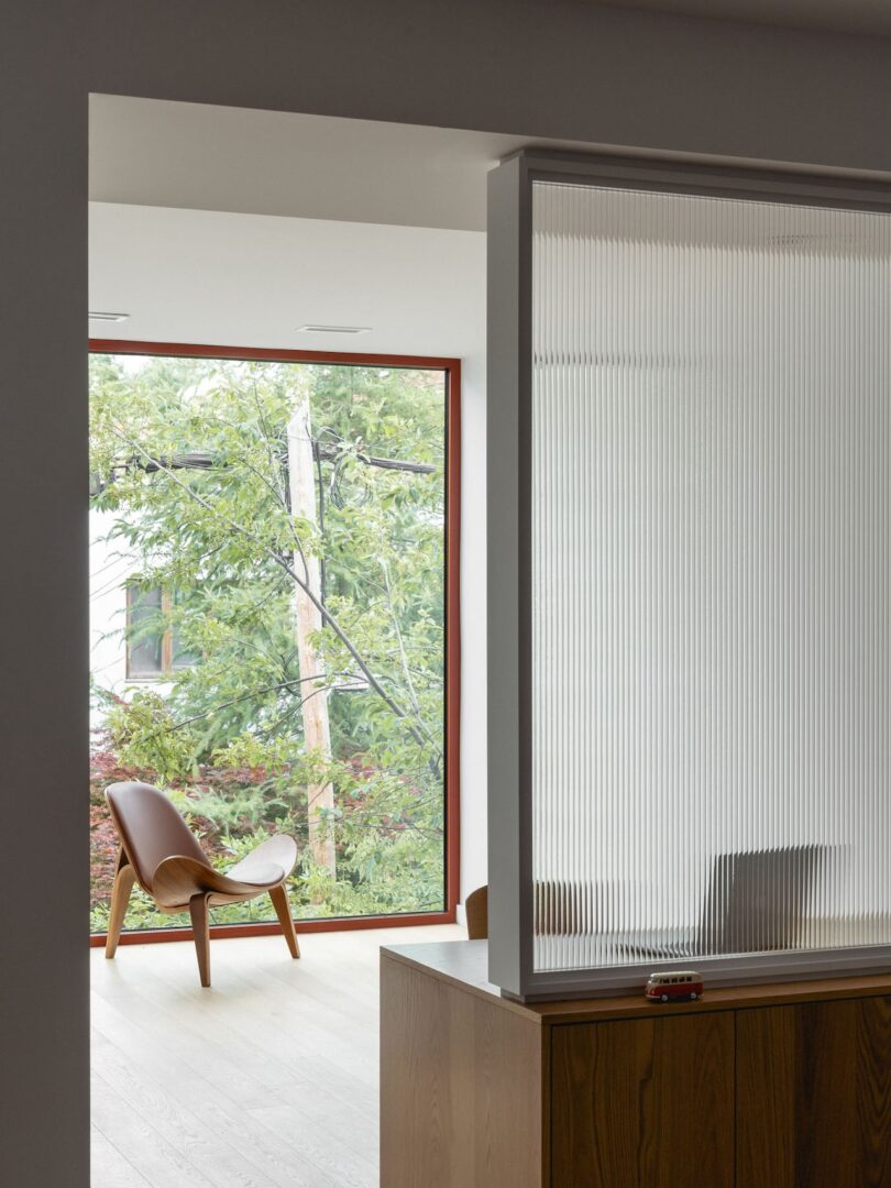 A modern interior features a wooden chair by a large window overlooking greenery. Fluted glass partition and wooden furniture are visible in the foreground.