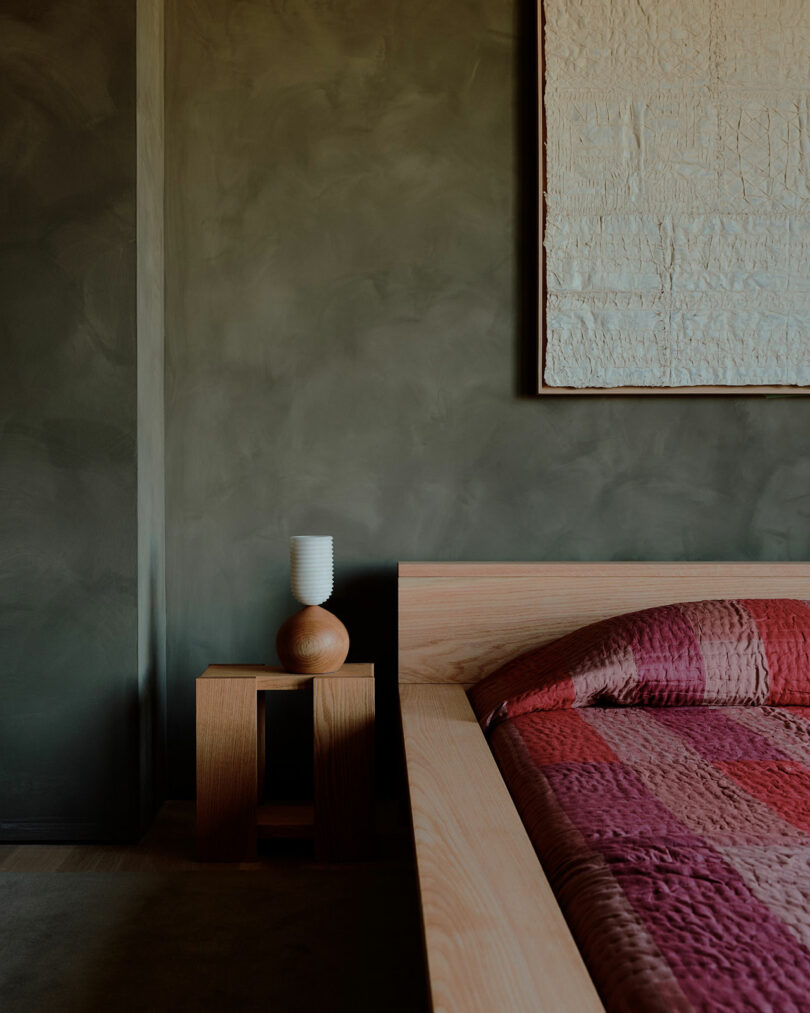 Bedroom corner with a wooden bed, red quilt, small wooden side table, and a white textured wall hanging against a dark olive wall.