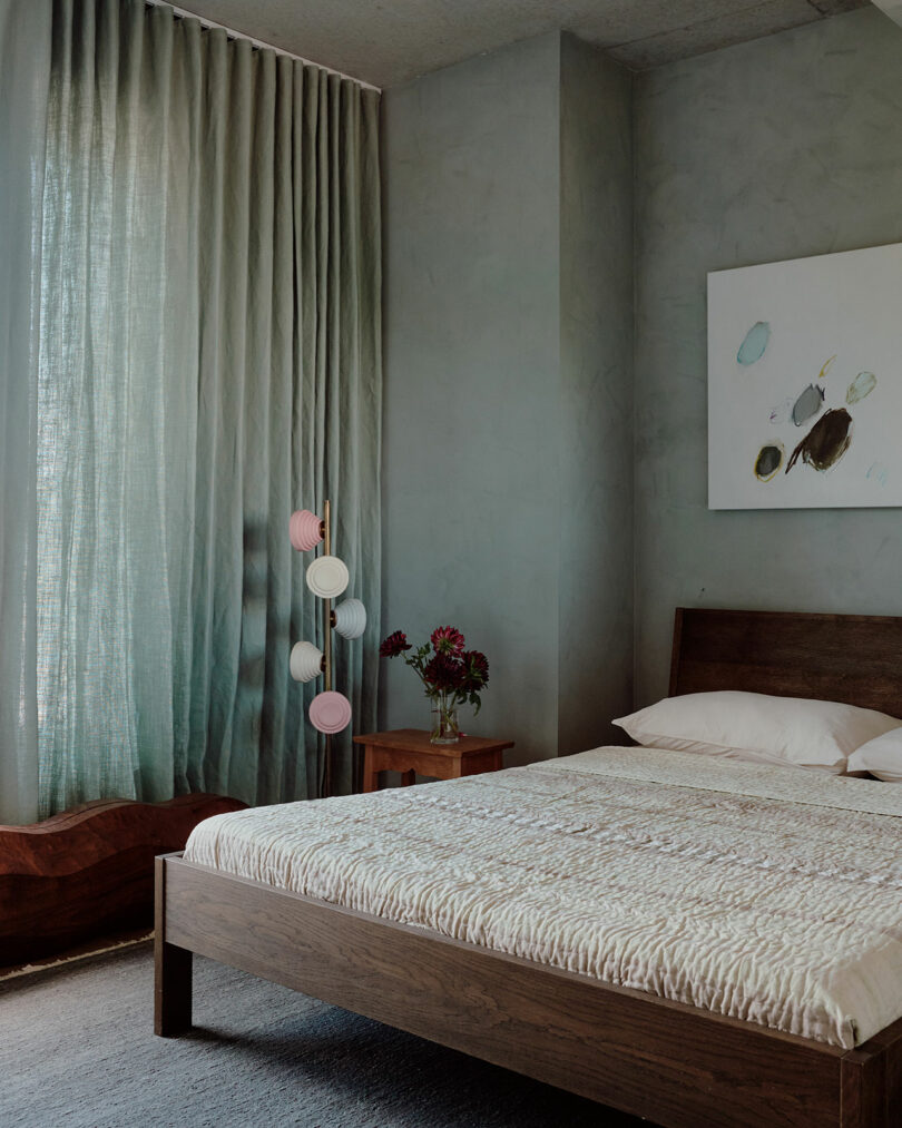 A bedroom with a wooden bed, textured bedding, bedside table with flowers, abstract art on the wall, and sheer curtains filtering soft light.