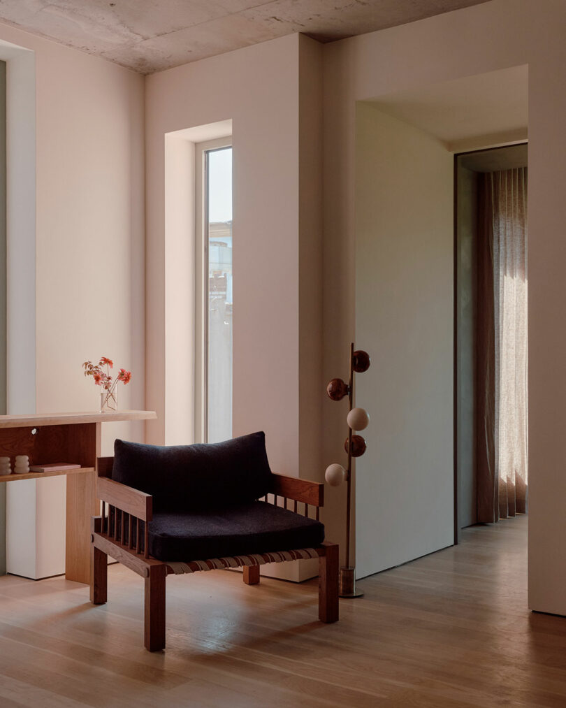 A minimalist room with a wooden chair, a small table holding a vase of flowers, and a floor lamp. Natural light comes through a window and an open door reveals a hallway with curtains.