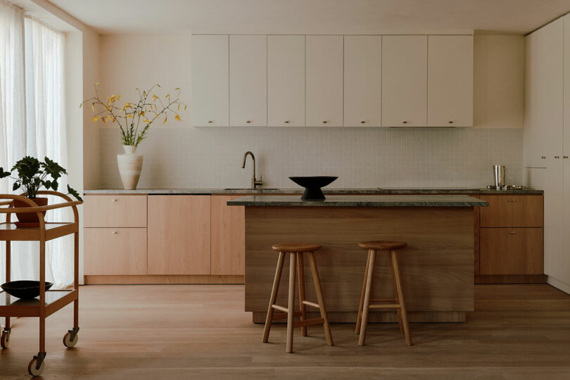 Minimalist kitchen with light wood cabinets, an island with two stools, a serving cart, and a vase with flowers on the counter.