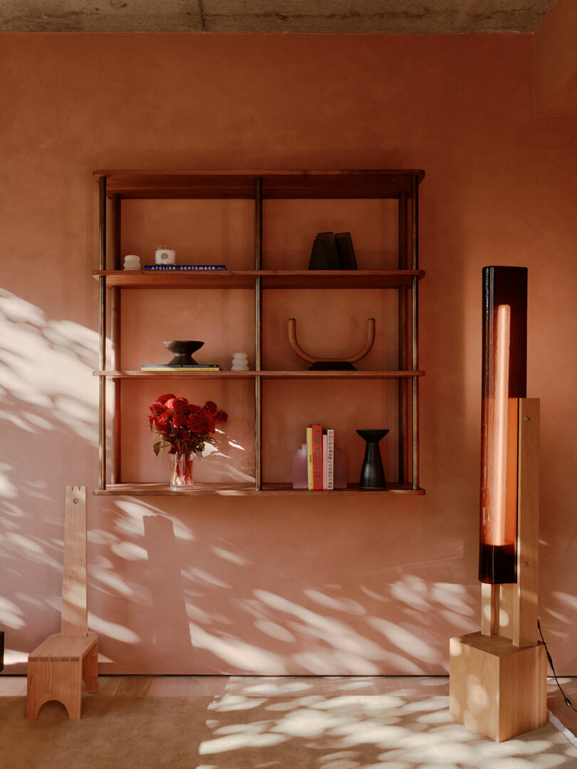 Room with pink walls, a wooden shelf with books and decor, a vase with red flowers, a wooden chair, and a tall lamp casting shadows.