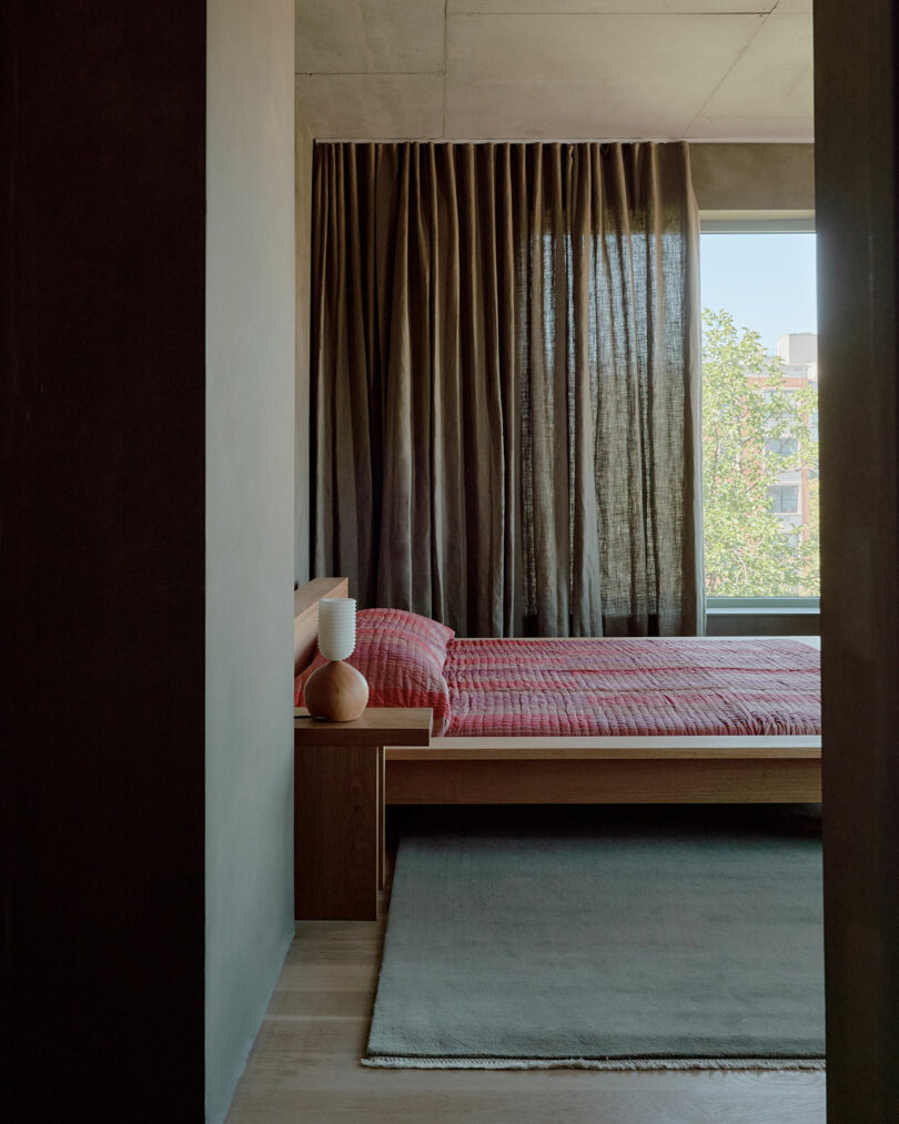 Minimalist bedroom with a wooden bed, red bedding, and a lamp on the nightstand. Dark curtains cover the window, and a muted rug lies on the floor.