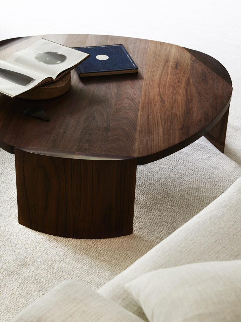 A round wooden coffee table with two books and a decorative item on top, placed on a light-colored rug next to a sofa.