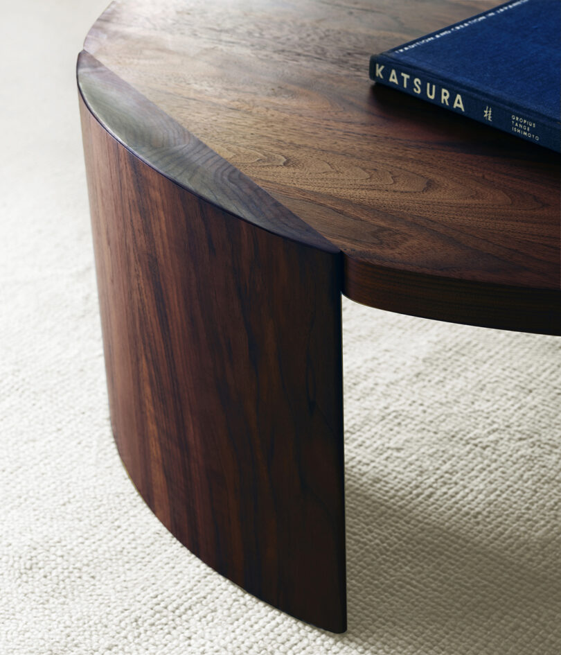 Close-up of a wooden coffee table with a curved support, resting on a textured carpet, and a blue book titled "Katsura" placed on top.