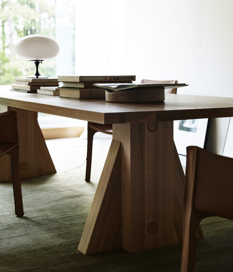 Wooden table with geometric legs, stacked books, and a lamp on top, surrounded by brown chairs. Large window in the background.