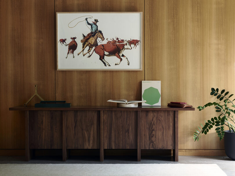 A wooden sideboard against a wood-paneled wall, featuring a cowboy artwork above it. A small sculpture, books, and a potted plant are displayed on the sideboard.