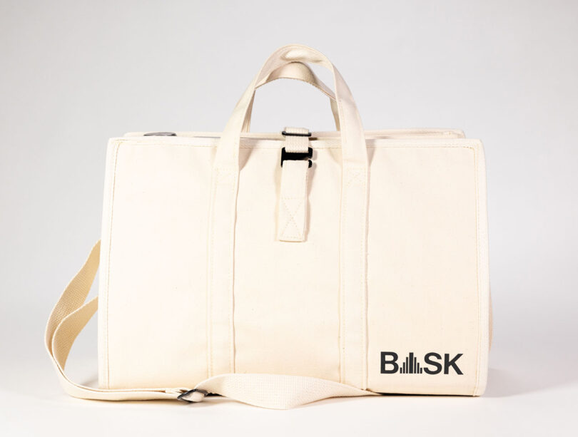 The BASK Bag from two-point is a beige canvas accessory with sturdy handles and a shoulder strap, featuring the iconic BASK collaboration logo prominently on the front.