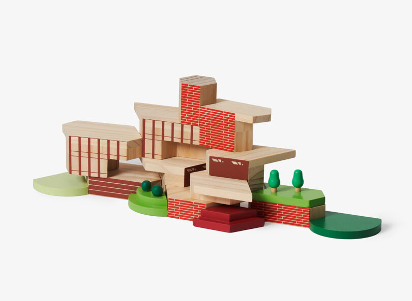 Wooden building blocks assembled to resemble a modern house design with red brick and natural wood patterns, surrounded by green pieces representing grass and trees.