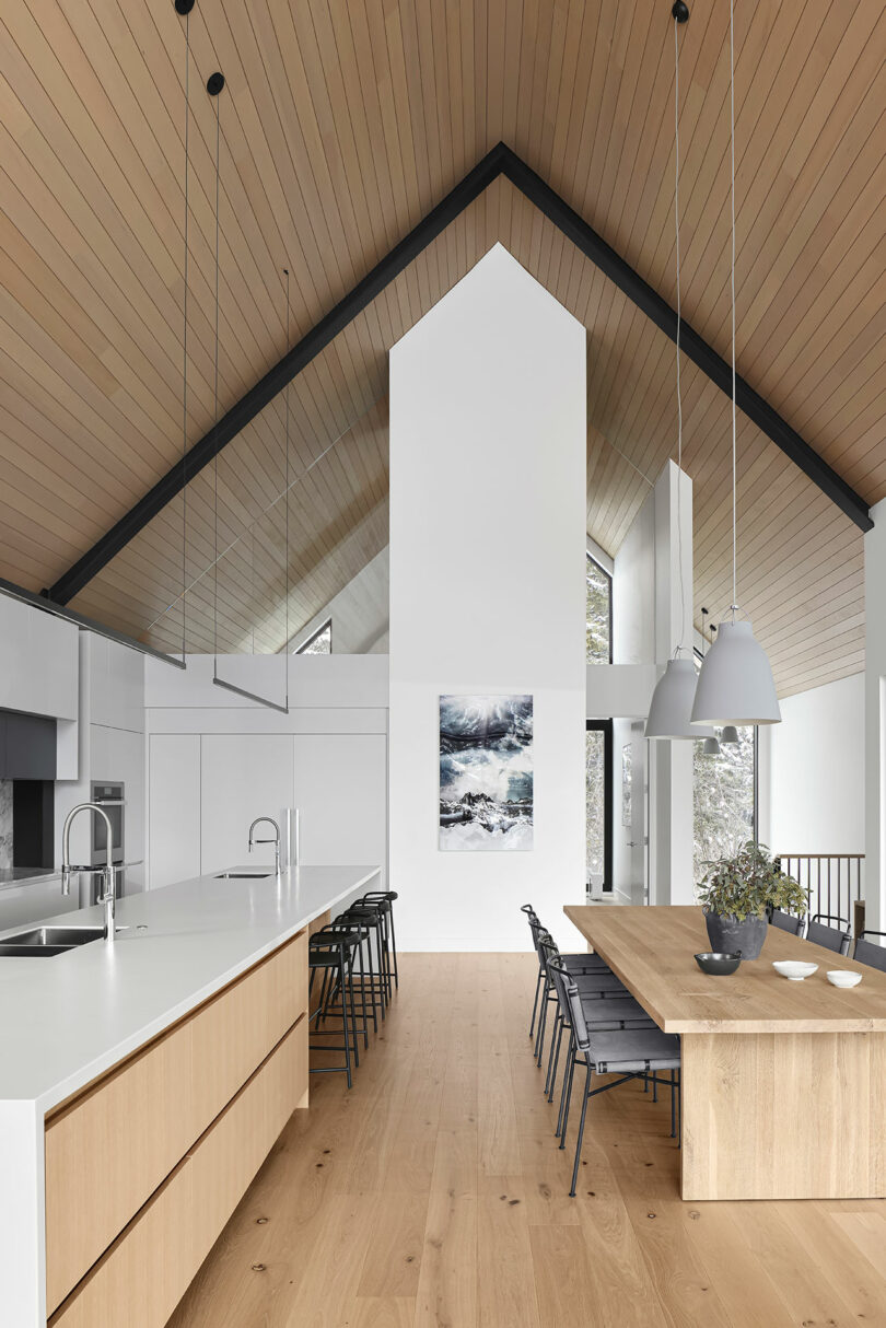 Modern kitchen and dining area with high wooden ceilings, a long island with stools, a wooden dining table with chairs, pendant lights, and a painting on a white wall.