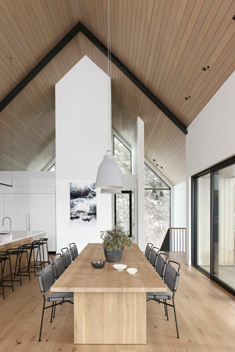 Modern dining area with a long wooden table, black chairs, pendant lighting, and large windows overlooking a snow-covered landscape.