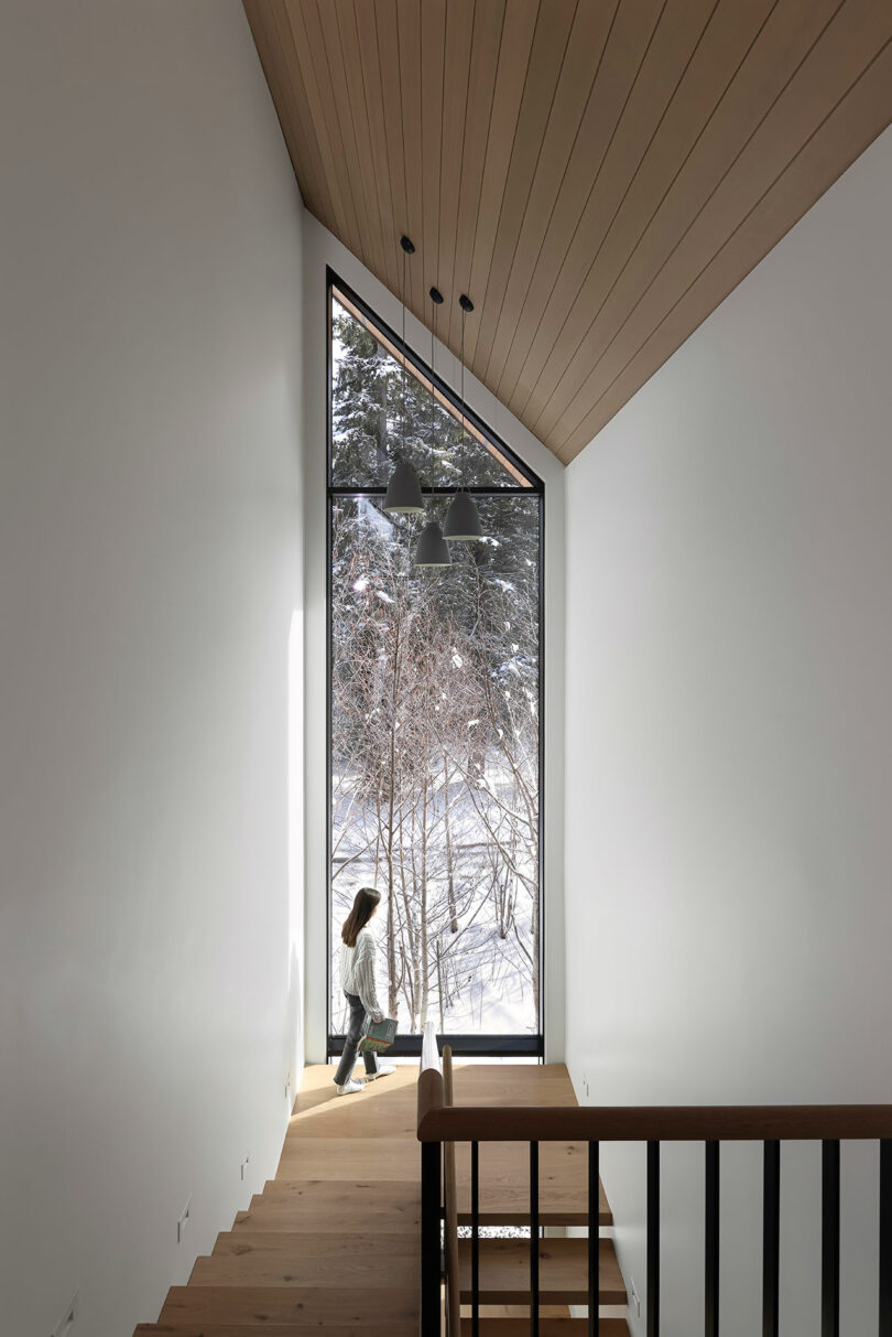 A person walking down a wooden staircase beside a large window with a snowy outdoor view.