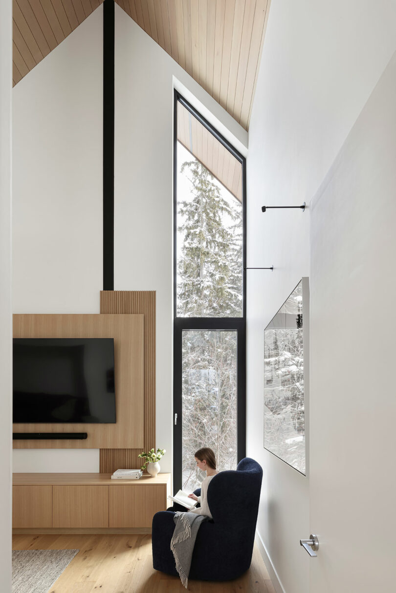 A person sitting in a black chair reads by a large window in a modern living room. The room features a wooden TV panel and has tall ceilings with wooden accents. Snowy trees are visible outside.