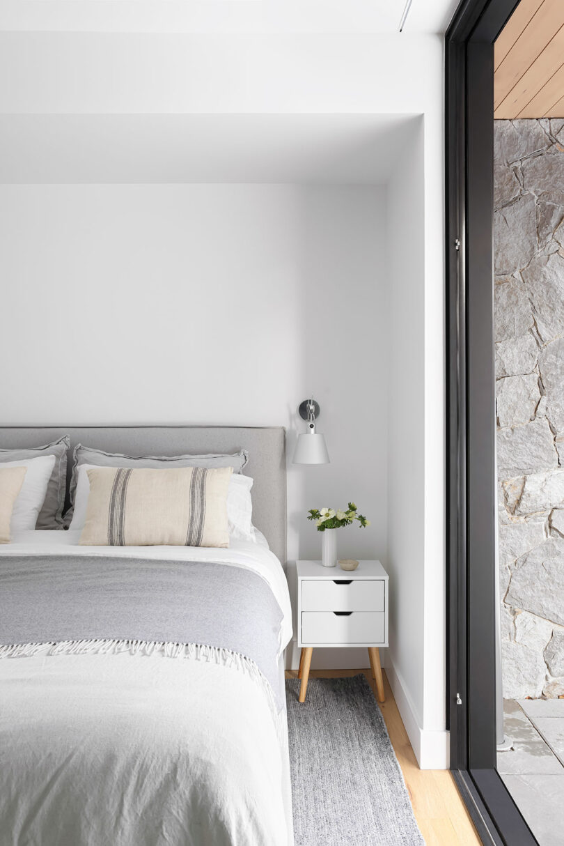 Minimalist bedroom with a bed, white and grey bedding, a bedside table with a lamp and plant, next to a large sliding glass door opening to a stone wall.